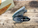 A pair of dark gray sneakers with yellow accents is placed on a rustic wooden surface. The sneakers have a modern, athletic design with visible treads on the soles. Nearby, a shoebox with the words 'MADE IN SPAIN' in red letters is partially visible.