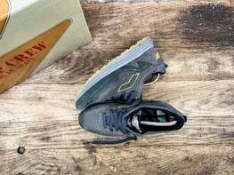 A pair of dark gray sneakers with yellow accents is placed on a rustic wooden surface. The sneakers have a modern, athletic design with visible treads on the soles. Nearby, a shoebox with the words 'MADE IN SPAIN' in red letters is partially visible.