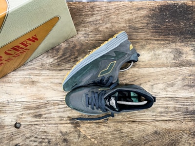 A pair of dark gray sneakers with yellow accents is placed on a rustic wooden surface. The sneakers have a modern, athletic design with visible treads on the soles. Nearby, a shoebox with the words 'MADE IN SPAIN' in red letters is partially visible.