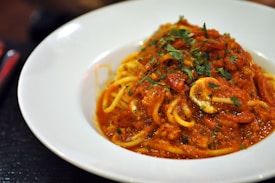 A plate of spaghetti topped with a rich red tomato sauce, garnished with fresh green herbs. The pasta appears to be cooked perfectly and the sauce looks thick, with visible tomato chunks and possibly slices of mushrooms.
