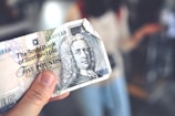 A hand holding a five-pound sterling banknote from the Royal Bank of Scotland, featuring intricate designs and the image of a historical figure with curly hair. The background is blurred, focusing attention on the banknote.