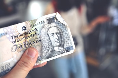 A hand holding a five-pound sterling banknote from the Royal Bank of Scotland, featuring intricate designs and the image of a historical figure with curly hair. The background is blurred, focusing attention on the banknote.