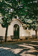 An ornate entrance labeled 'Casal de S. Pedro', adorned with a decorative archway and tiled details. It is surrounded by lush green trees casting shadows on the cobblestone pathway with benches placed alongside.