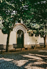 An ornate entrance labeled 'Casal de S. Pedro', adorned with a decorative archway and tiled details. It is surrounded by lush green trees casting shadows on the cobblestone pathway with benches placed alongside.
