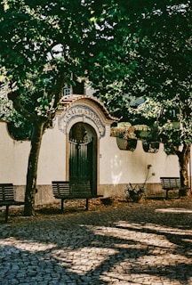 An ornate entrance labeled 'Casal de S. Pedro', adorned with a decorative archway and tiled details. It is surrounded by lush green trees casting shadows on the cobblestone pathway with benches placed alongside.