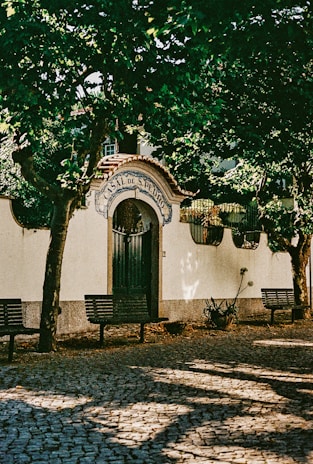 An ornate entrance labeled 'Casal de S. Pedro', adorned with a decorative archway and tiled details. It is surrounded by lush green trees casting shadows on the cobblestone pathway with benches placed alongside.