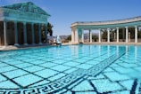 A large, luxurious swimming pool with clear, blue water surrounded by classical architecture, including columns and statues. The pool has an intricate tile pattern on the bottom, and there are hills visible in the background under a bright, clear sky.