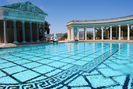 A large, luxurious swimming pool with clear, blue water surrounded by classical architecture, including columns and statues. The pool has an intricate tile pattern on the bottom, and there are hills visible in the background under a bright, clear sky.