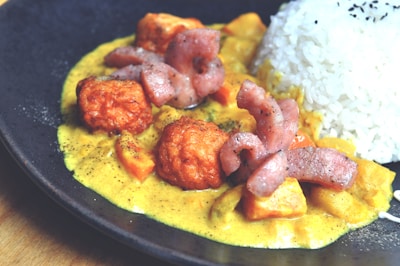 A plate of food featuring creamy curry sauce with diced vegetables, golden-brown fried chicken pieces, and strips of seared meat. On the side, there is a portion of steamed white rice topped with black sesame seeds. The dish is artistically plated on a dark, round plate.