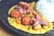 A plate of food featuring creamy curry sauce with diced vegetables, golden-brown fried chicken pieces, and strips of seared meat. On the side, there is a portion of steamed white rice topped with black sesame seeds. The dish is artistically plated on a dark, round plate.