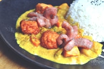 A plate of food featuring creamy curry sauce with diced vegetables, golden-brown fried chicken pieces, and strips of seared meat. On the side, there is a portion of steamed white rice topped with black sesame seeds. The dish is artistically plated on a dark, round plate.
