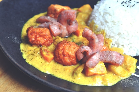 A plate of food featuring creamy curry sauce with diced vegetables, golden-brown fried chicken pieces, and strips of seared meat. On the side, there is a portion of steamed white rice topped with black sesame seeds. The dish is artistically plated on a dark, round plate.