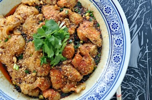 A dish featuring tender slices of meat coated in a flavorful mixture of spices and sesame seeds. Garnished with fresh cilantro, the dish is served in a white and blue patterned bowl placed on a dark textured surface.