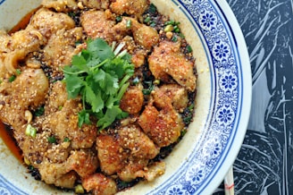 A dish featuring tender slices of meat coated in a flavorful mixture of spices and sesame seeds. Garnished with fresh cilantro, the dish is served in a white and blue patterned bowl placed on a dark textured surface.