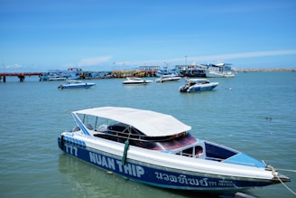 Numerous speedboats and other small watercraft are anchored or floating in a calm harbor on a bright, sunny day. The nearest speedboat has blue and white hull with a canopy, featuring the text 'NUAN THIP' on its side. The background showcases a vibrant blue sky, a long pier, and additional boats further in the distance.