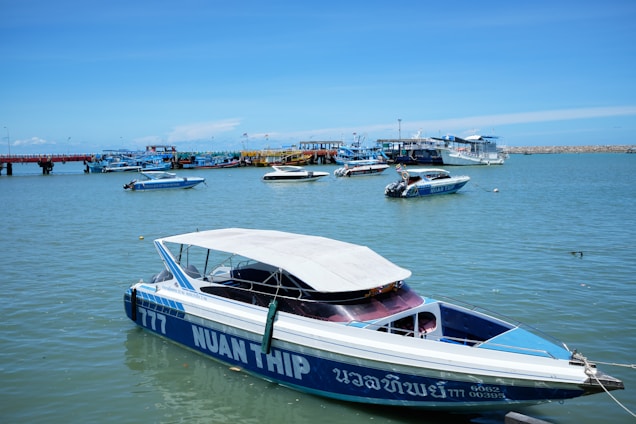 Numerous speedboats and other small watercraft are anchored or floating in a calm harbor on a bright, sunny day. The nearest speedboat has blue and white hull with a canopy, featuring the text 'NUAN THIP' on its side. The background showcases a vibrant blue sky, a long pier, and additional boats further in the distance.