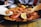 A large pan filled with seafood paella, showcasing prawns, mussels, and a slice of lemon on top of rice infused with a rich, golden color. Some greens appear mixed within the rice, and the scene includes a close-up view of the dish with a blurred background of a person in a plaid shirt sitting at the table.