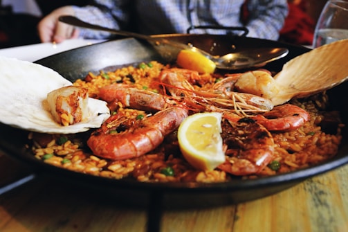 A large pan filled with seafood paella, showcasing prawns, mussels, and a slice of lemon on top of rice infused with a rich, golden color. Some greens appear mixed within the rice, and the scene includes a close-up view of the dish with a blurred background of a person in a plaid shirt sitting at the table.