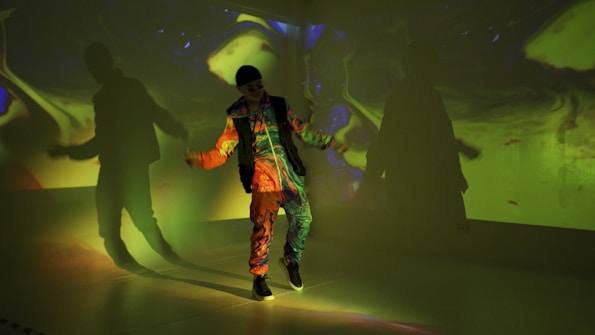 A person in colorful, vibrant clothing dances with expressive hand gestures. The room is filled with psychedelic, abstract light projections on the walls, casting dramatic shadows and creating a surreal atmosphere.