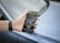 A hand holds a smartphone displaying a map and navigation interface, with points of interest highlighted. The phone is held by someone wearing a dark wristband. In the background, there is a blurred view of a car surface.