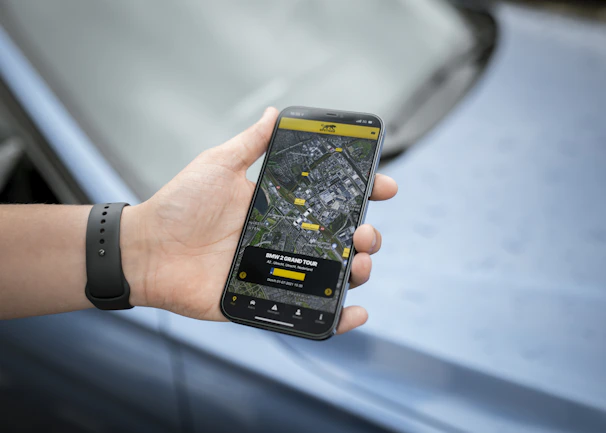 A hand holds a smartphone displaying a map and navigation interface, with points of interest highlighted. The phone is held by someone wearing a dark wristband. In the background, there is a blurred view of a car surface.