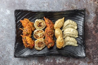 An assortment of dumplings is neatly arranged on a rectangular black plate. The dish includes fried, steamed, and possibly pan-seared dumplings with varying textures and colors, ranging from golden brown to light beige.