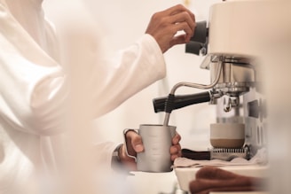 A person in a white garment is using a coffee machine, with one hand adjusting a control and the other holding a stainless steel frothing pitcher under the steam wand. A cup with a white and brown design sits nearby, ready to be filled.