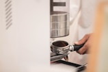 A hand holds a portafilter filled with ground coffee, positioned beneath a coffee grinder. The scene suggests a process of coffee preparation, with focus on the equipment used for making espresso.