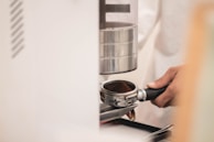 A hand holds a portafilter filled with ground coffee, positioned beneath a coffee grinder. The scene suggests a process of coffee preparation, with focus on the equipment used for making espresso.