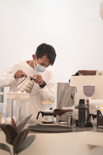 A person wearing a face mask is carefully pouring hot water from a kettle into a coffee dripper over a glass carafe on a countertop. Various coffee-making tools and appliances are arranged on the counter, including a grinder and a scale. The setting appears calm and focused, likely in a coffee shop or home brewing setup.