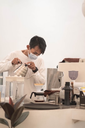 A person wearing a face mask is carefully pouring hot water from a kettle into a coffee dripper over a glass carafe on a countertop. Various coffee-making tools and appliances are arranged on the counter, including a grinder and a scale. The setting appears calm and focused, likely in a coffee shop or home brewing setup.