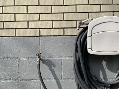 A neatly coiled black garden hose hangs on a light gray holder attached to a brick wall. The wall is composed of yellowish bricks on the top section and painted cement blocks on the lower section. An outdoor water spigot is visible on the left side of the image, casting a shadow.