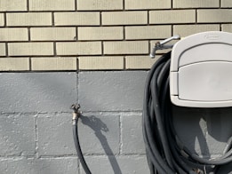 A neatly coiled black garden hose hangs on a light gray holder attached to a brick wall. The wall is composed of yellowish bricks on the top section and painted cement blocks on the lower section. An outdoor water spigot is visible on the left side of the image, casting a shadow.