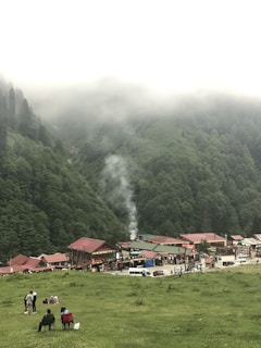 A small village nestled at the base of a lush green mountain range with mist covering the peaks. The village has several buildings with red roofs and a few vehicles parked along the street. People are gathered on a grassy area in the foreground, some sitting on chairs while others stand or are engaged in conversation. A plume of smoke rises from one of the buildings, adding to the tranquil atmosphere.