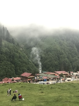 A small village nestled at the base of a lush green mountain range with mist covering the peaks. The village has several buildings with red roofs and a few vehicles parked along the street. People are gathered on a grassy area in the foreground, some sitting on chairs while others stand or are engaged in conversation. A plume of smoke rises from one of the buildings, adding to the tranquil atmosphere.
