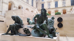 Bronze statues are prominently displayed in a museum gallery with classical architecture. The sculptures depict dramatic and expressive human figures in various poses. The space features arches and rectangular stone walls with decorative elements, and natural light streams in from the windows above.