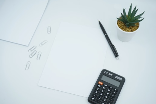 A minimalist workspace setup featuring a black calculator, a black pen, and a small white potted plant with green leaves. There are blank sheets of paper and a few metal paperclips scattered on a white surface.