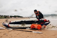 A person in a black wetsuit with an orange life vest is attending to an inflatable kayak on a sandy beach. The kayak is labeled 'BARAM 200' and carries orange and red gear. In the background, there is a coastline with houses and hills under a cloudy sky, and the sea gently waves onto the shore.