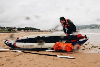 A person in a black wetsuit with an orange life vest is attending to an inflatable kayak on a sandy beach. The kayak is labeled 'BARAM 200' and carries orange and red gear. In the background, there is a coastline with houses and hills under a cloudy sky, and the sea gently waves onto the shore.