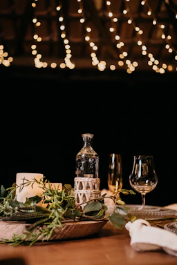 A cozy garden party table set with pink crystal wine glasses, gingham tablecloth, and soft glowing lights under a chandelier.