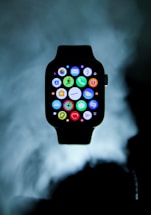 An Apple Watch is prominently displayed with its user interface showing numerous colorful app icons. The watch is set against a cloudy, dark background, giving it a floating appearance.