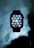 An Apple Watch is prominently displayed with its user interface showing numerous colorful app icons. The watch is set against a cloudy, dark background, giving it a floating appearance.