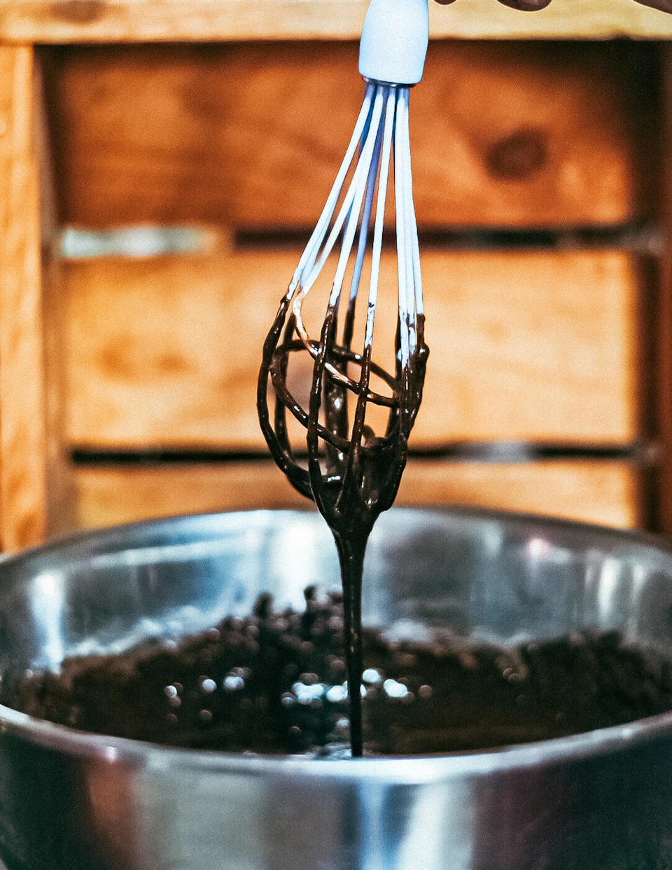 The image shows a silver mixing bowl filled with chocolate, being whisked by a white wire whisk held above it. The chocolate appears to be melted and smooth, indicating that it has been recently incorporated into the mixture. The background is blurred, drawing focus to the main subject of the scene - the whisk in motion stirring the chocolate within the bowl.

The image does not contain any discernible text or additional objects, with the primary focus being on the whisk and its interaction with the chocolate.
