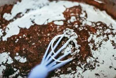 A close-up view of a whisk mixing dry cocoa powder with flour, creating a rustic, textured surface. The mixture appears partially combined, with visible clumps and streaks of unmixed powder. The background shows a blurred, earthy color palette.