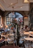 A luxurious dining room featuring a beautifully set table with wine glasses, a centerpiece of flowers, and napkins. The background includes a large arched window allowing soft natural light to fill the room, and there are subtle reflections on a shiny table surface.