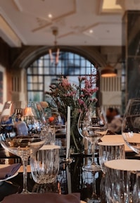 A luxurious dining room featuring a beautifully set table with wine glasses, a centerpiece of flowers, and napkins. The background includes a large arched window allowing soft natural light to fill the room, and there are subtle reflections on a shiny table surface.