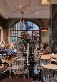 A luxurious dining room featuring a beautifully set table with wine glasses, a centerpiece of flowers, and napkins. The background includes a large arched window allowing soft natural light to fill the room, and there are subtle reflections on a shiny table surface.