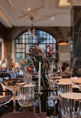 A luxurious dining room featuring a beautifully set table with wine glasses, a centerpiece of flowers, and napkins. The background includes a large arched window allowing soft natural light to fill the room, and there are subtle reflections on a shiny table surface.