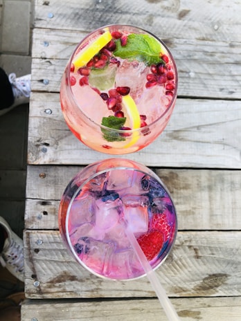 Two refreshing beverages placed on a rustic wooden table. The upper glass contains a pink drink with pomegranate seeds, lemon slices, and mint leaves. The lower glass holds a clear drink with pink hues and visible ice cubes, garnished with a strawberry.