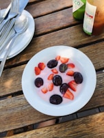 A bowl containing pink yogurt topped with plump dark grapes and sliced strawberries, placed on a wooden table. Nearby, there is a stack of plates with cutlery and a bottle of Tabasco sauce.